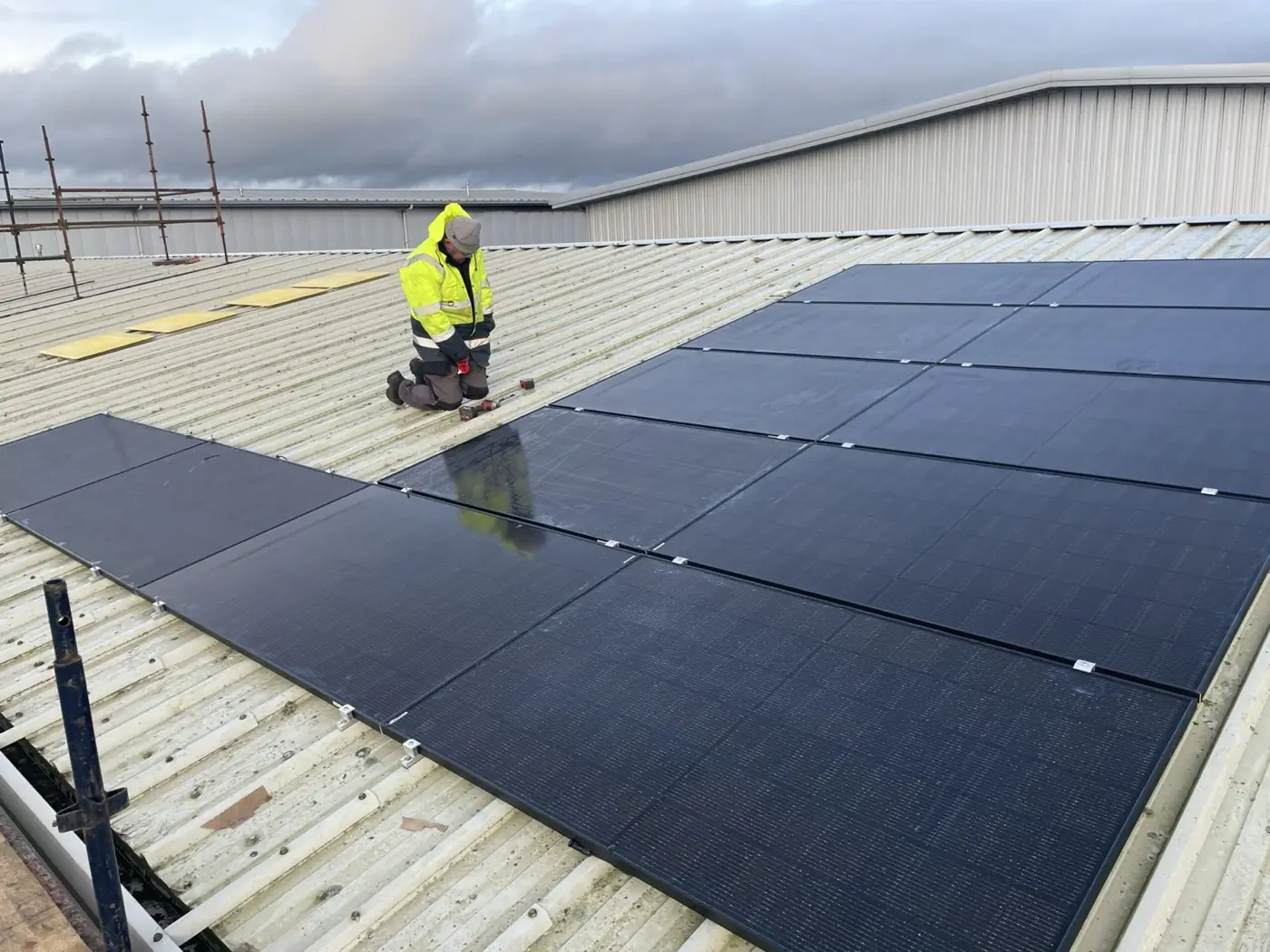 An installer in hi-vis kneeling on a metal roof, fitting a row of black solar panels
