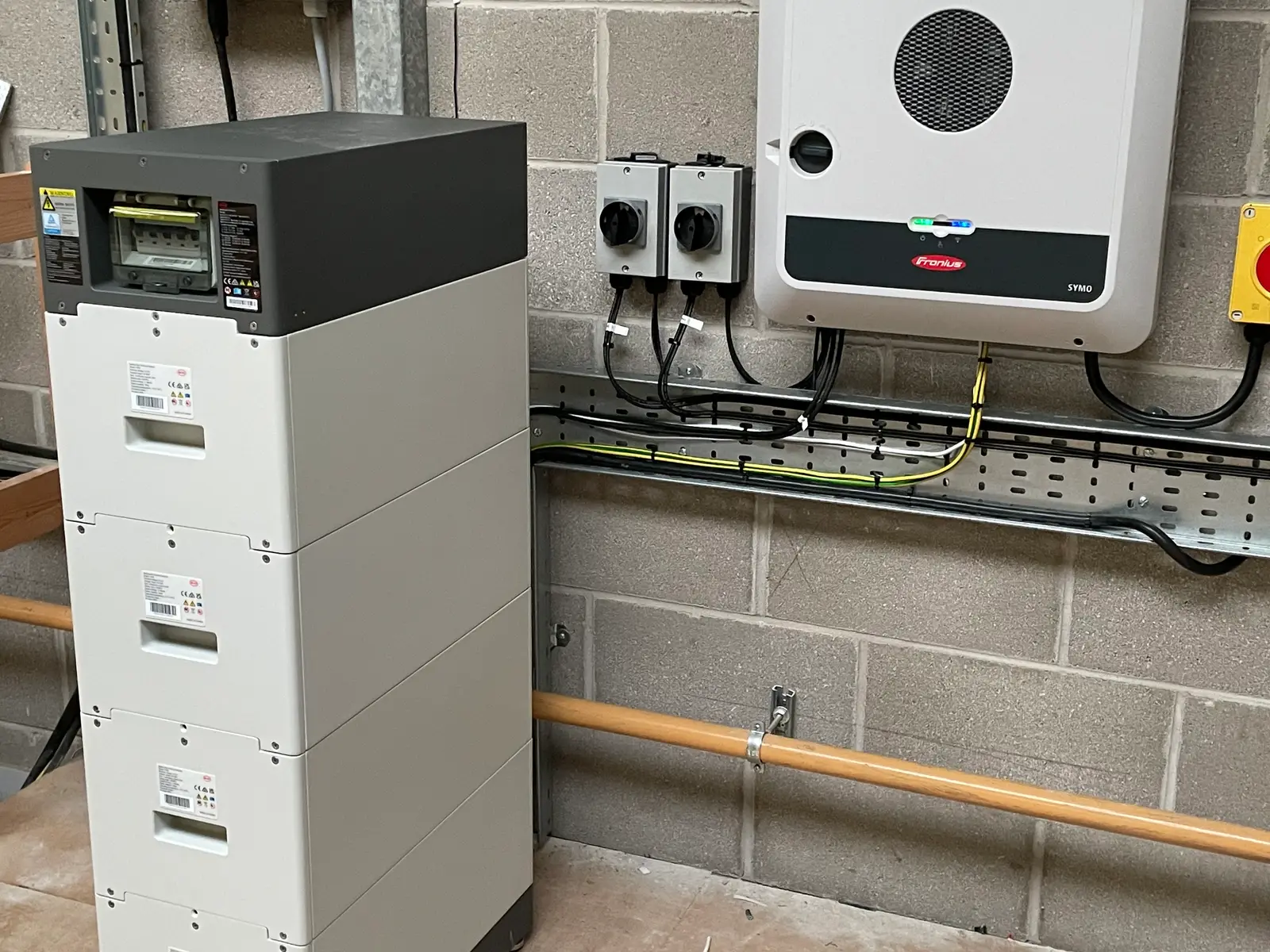 A Fronius hybrid inverter on a block wall, with a white BYD battery stack standing beside it