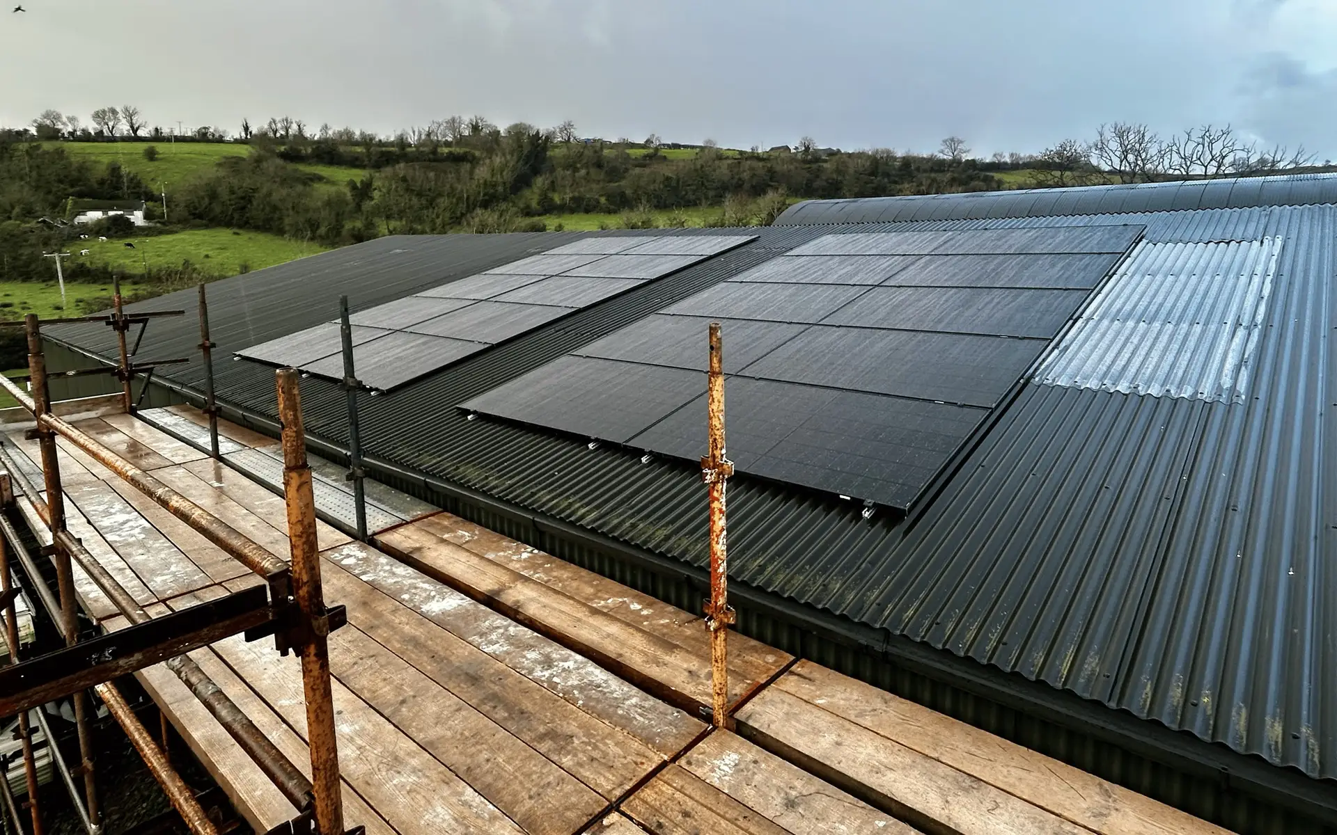 Black solar panels on a dark corrugated farm roof, seen from a scaffold with green hills behind